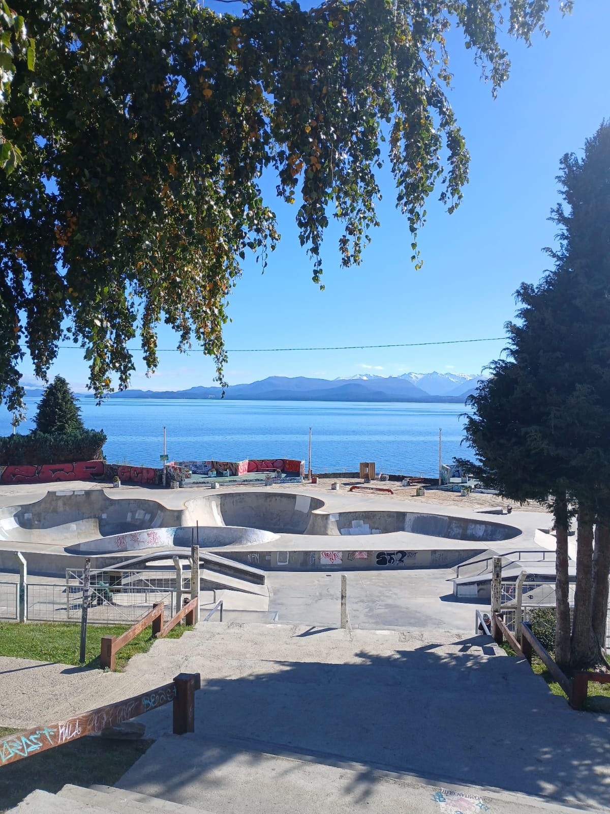 Vista del skatepark Baripark de Bariloche con sus bowls de cemento, el lago Nahuel Huapi y la cordillera de fondo, en un día soleado