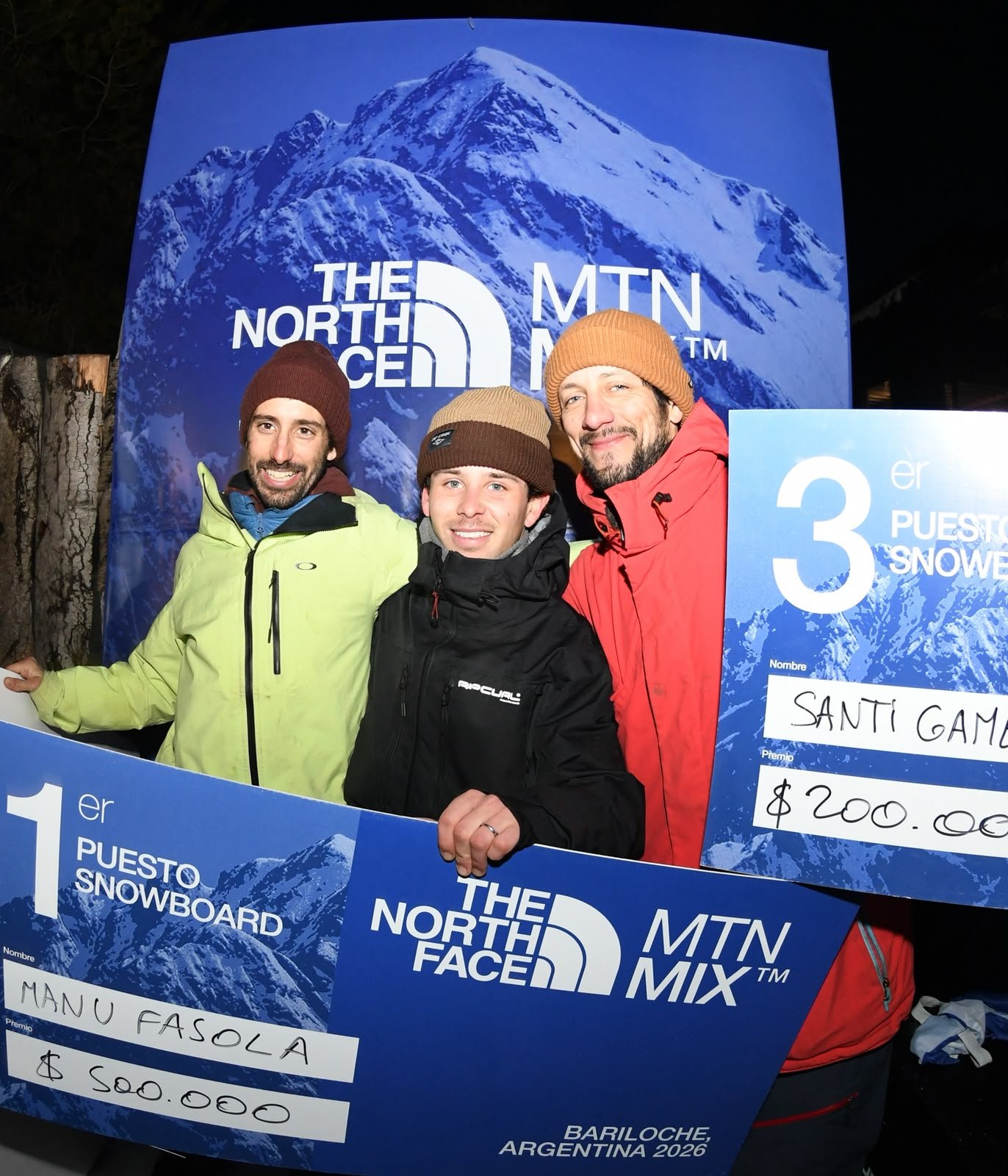 Los tres primeros puestos de snowboard del TNF Rail Jam posan con los cheques de premio, con el cartel de Bariloche, Argentina 2026 de fondo